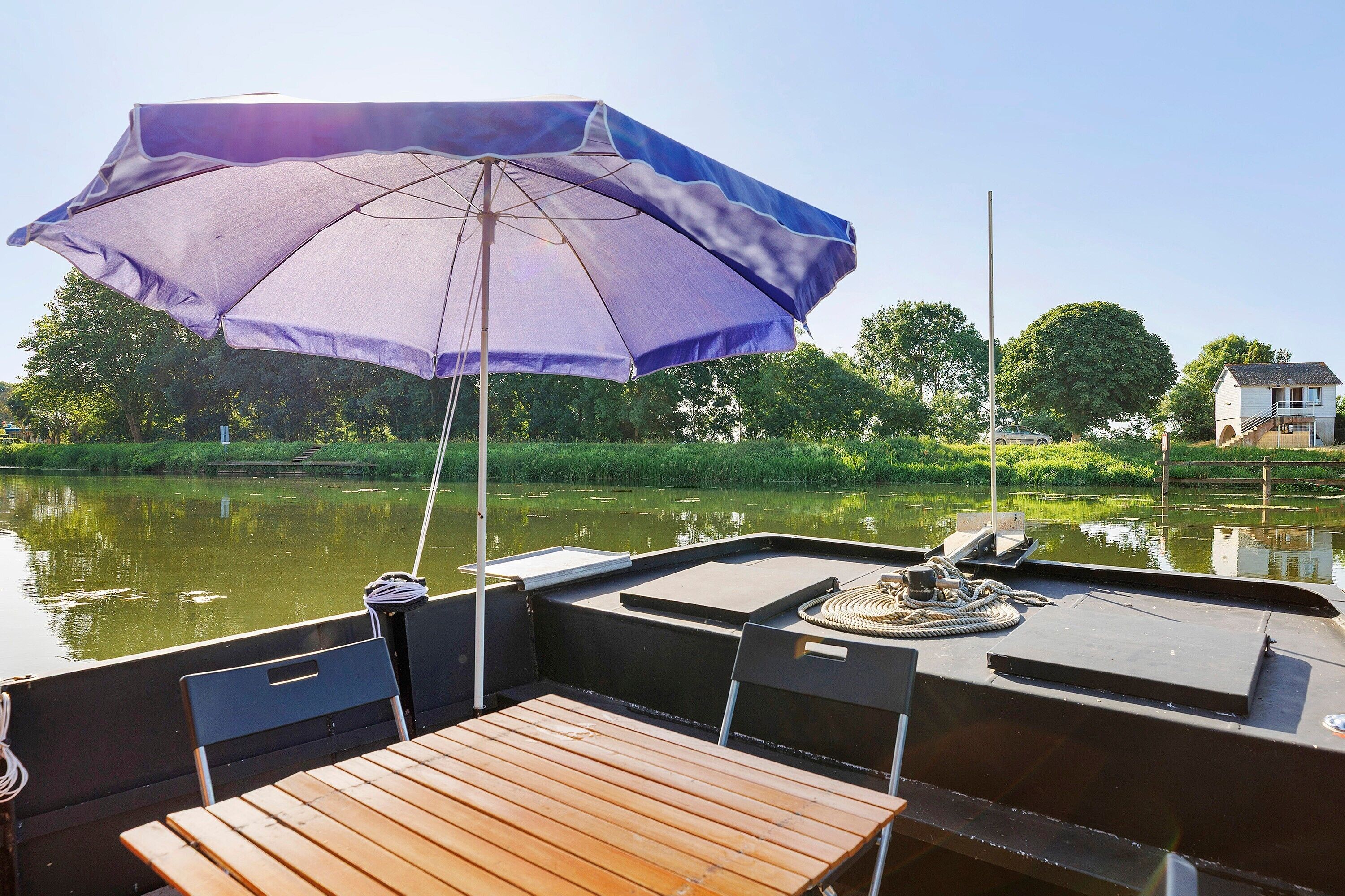 Un bateau avec une table et des chaises, un parasol, et une rivière paisible bordée de verdure, avec une maison en arrière-plan.