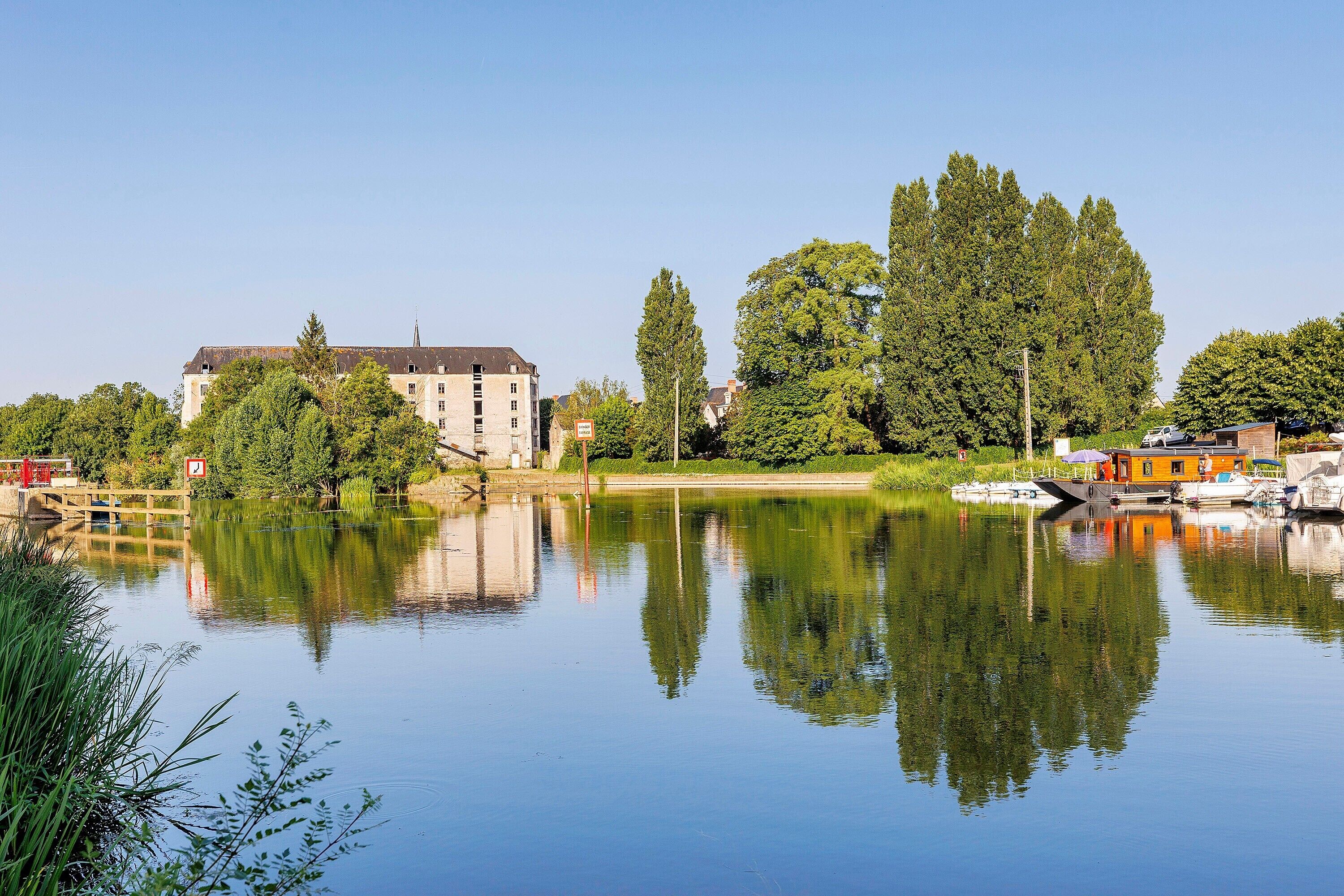 Un lac paisible où se reflètent les arbres et les bâtiments, un pont en bois et des bateaux amarrés près d’un bâtiment doté d’un toit en pente.