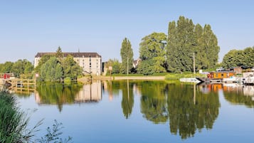 Un lac paisible où se reflètent les arbres et les bâtiments, un pont en bois et des bateaux amarrés près d’un bâtiment doté d’un toit en pente.