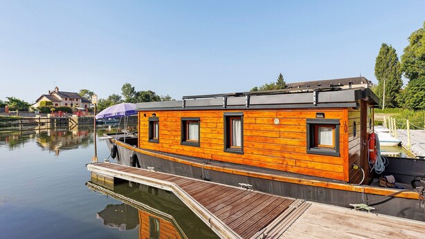 A wooden houseboat docked at a calm water body with a dock, a lifebuoy, and a small boat nearby.