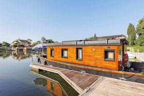 A wooden houseboat docked at a calm water body with a dock, a lifebuoy, and a small boat nearby.