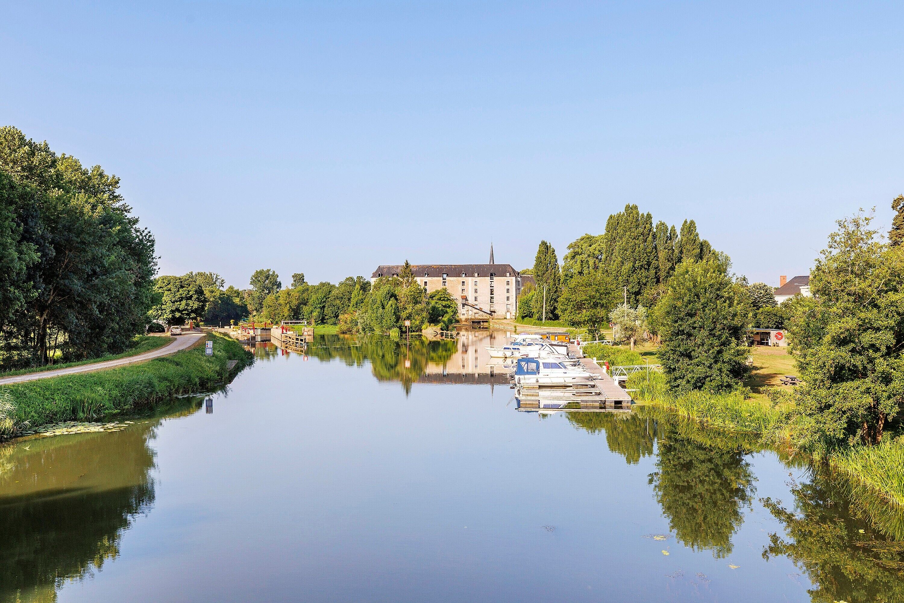 A calm river with boats docked, surrounded by lush greenery and a large building with a tower.