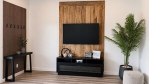 A modern living room with a flat-screen TV mounted on a wooden wall, a black entertainment unit below, and a potted plant to the right.