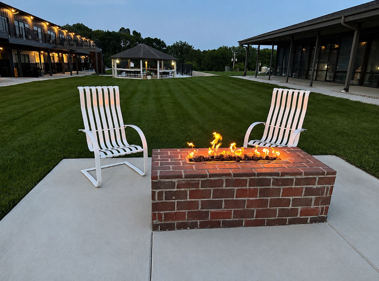 Un espace patio avec deux chaises Adirondack blanches, un foyer en briques où brûle un feu, et un gazebo en arrière-plan.