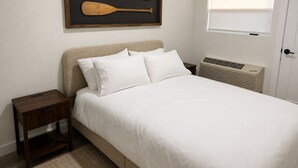 A bedroom with a bed, a nightstand, a wall-mounted picture frame, a window, and a radiator.
