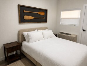Une chambre à coucher comprenant un lit, une table de chevet, un cadre photo fixé au mur, une fenêtre et un radiateur.