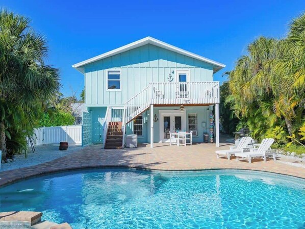 A light blue house with a balcony, a swimming pool, and a patio area with lounge chairs.