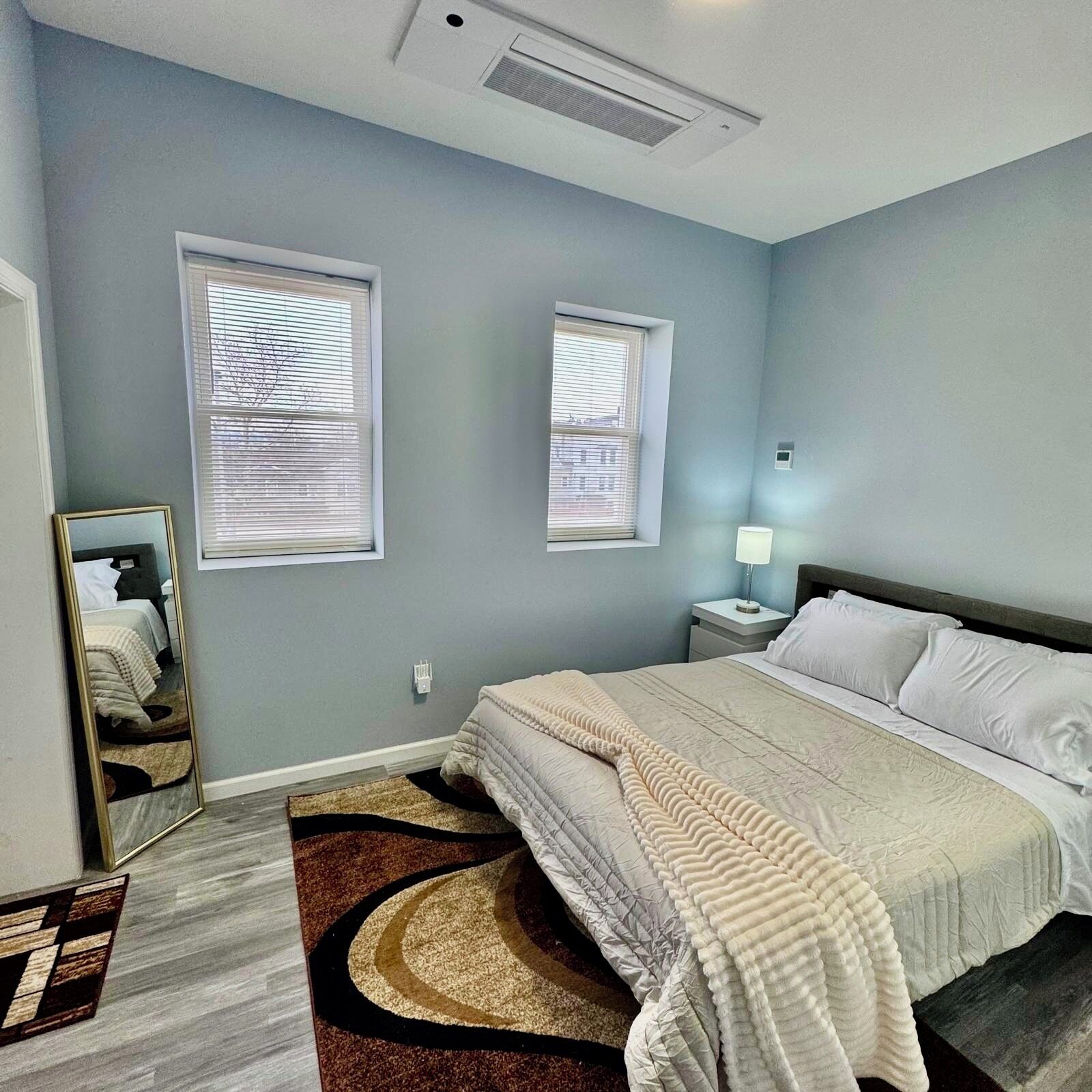 A bedroom with a bed, two windows, a nightstand, a mirror, and a rug.
