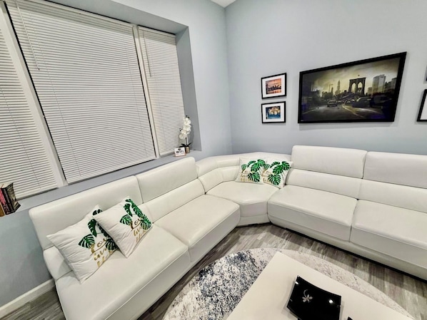 Un salon moderne avec un canapé d’angle blanc, une table basse ronde et un tableau représentant un paysage urbain accroché au mur.