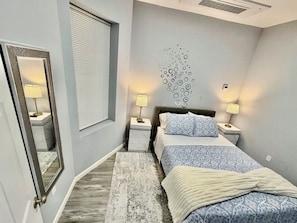 Ein Schlafzimmer mit Bett, Nachttischlampen, einem Spiegel, einem Fenster mit Jalousien und einem Wandbild.
