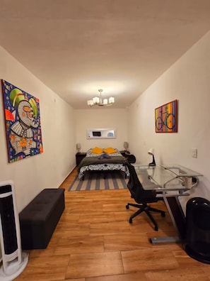 Ein schmales Zimmer mit einem Bett, einem Schreibtisch mit Stuhl, einem Heizkörper und Bildern an der Wand.