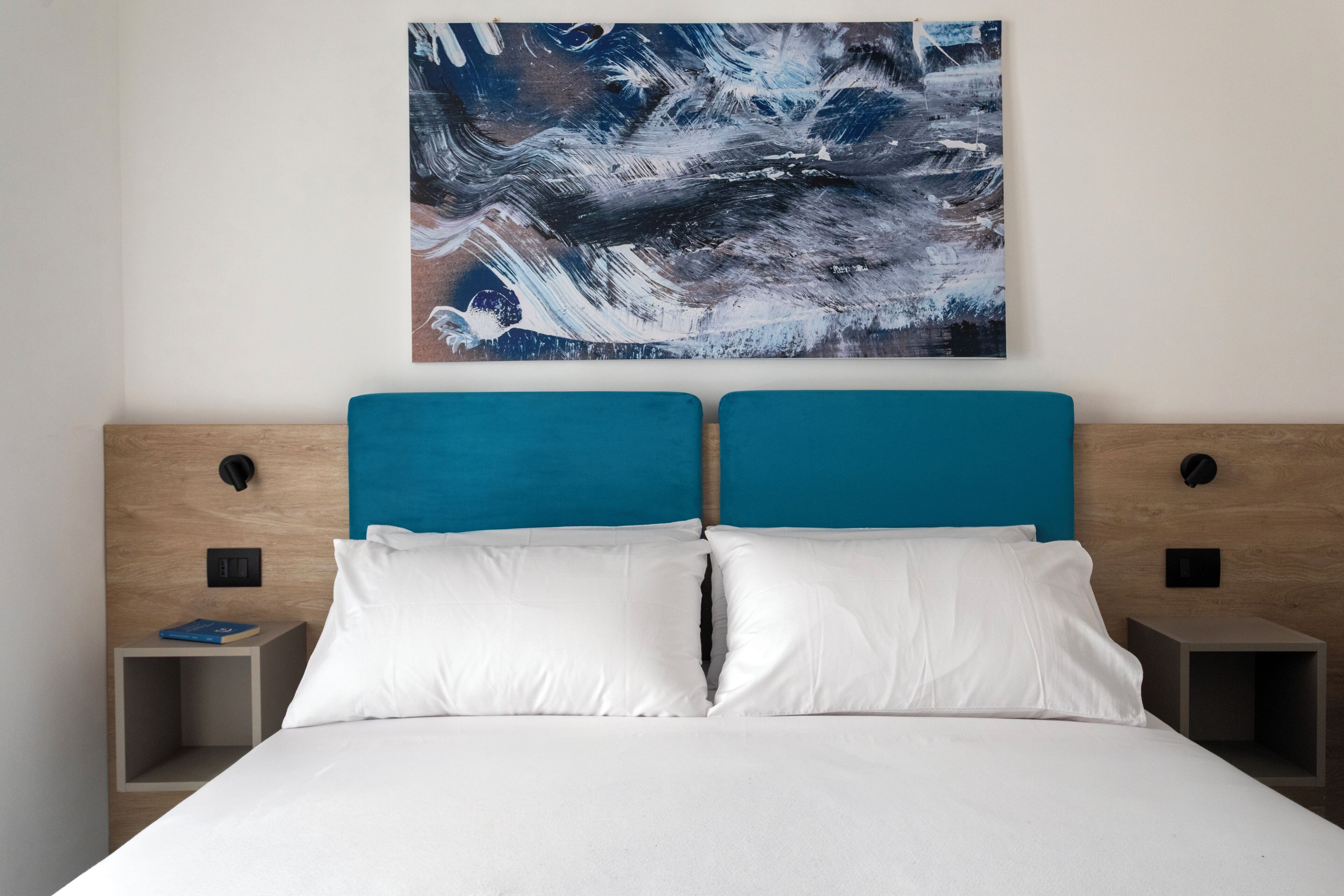 Ein modernes Schlafzimmer mit blauem Kopfteil, weißer Bettwäsche und einem großen abstrakten Gemälde an der Wand.
