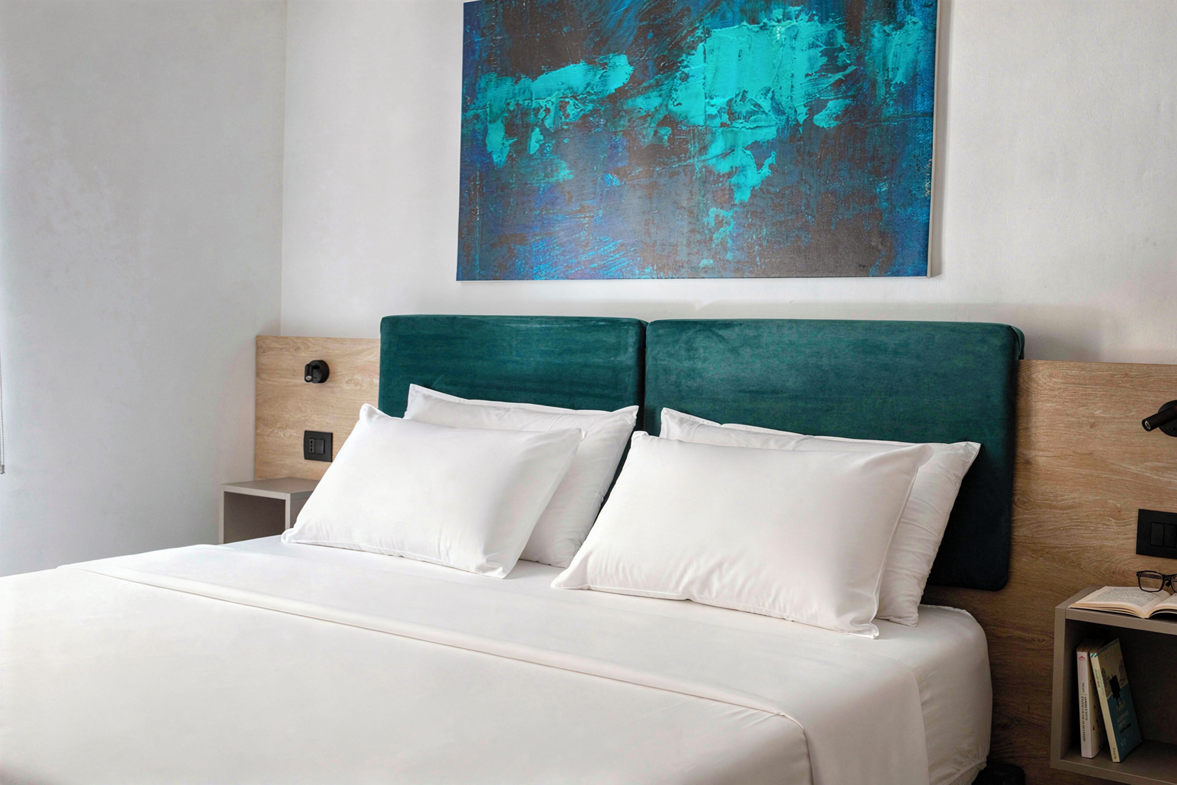 A neatly made bed with white bedding and a teal headboard, a small bedside table with a book, and a wall-mounted abstract painting.