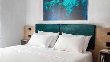A neatly made bed with white bedding and a teal headboard, a small bedside table with a book, and a wall-mounted abstract painting.