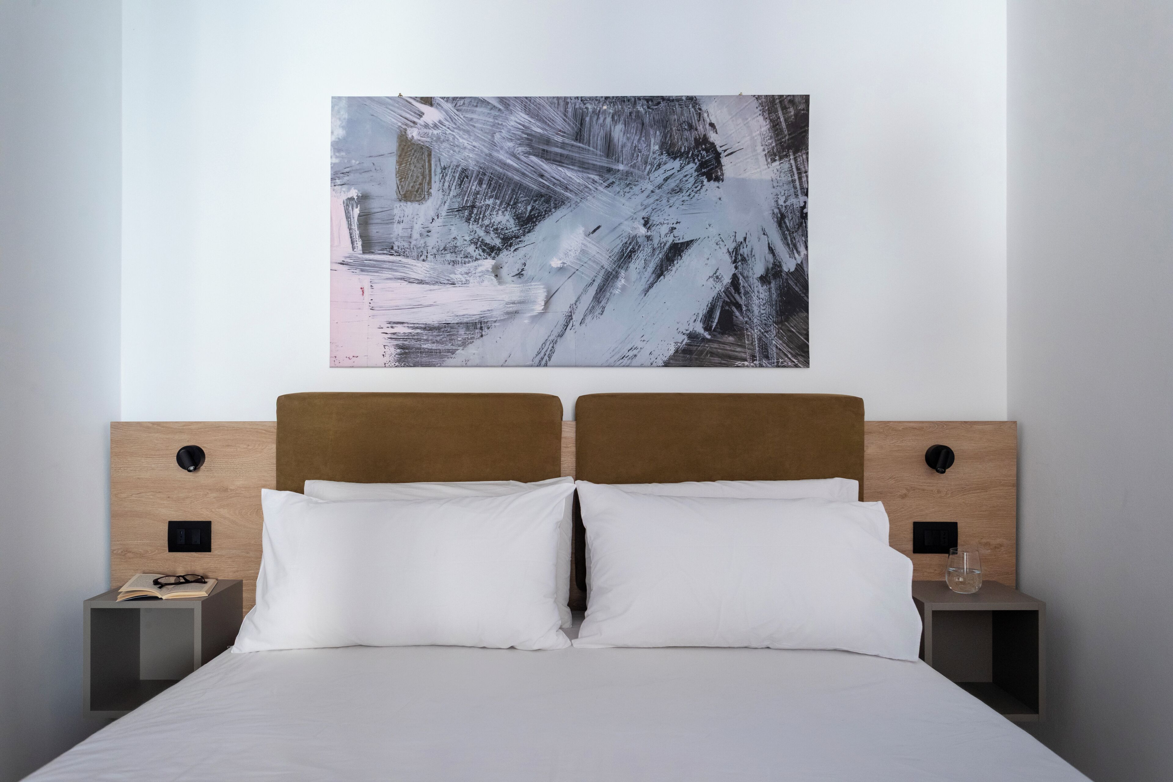 A modern hotel room with a large abstract painting, a wooden headboard, two bedside tables, and a neatly made bed.