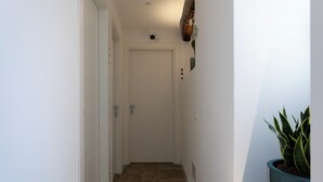 Un pasillo estrecho con una puerta blanca, una planta en maceta y una placa numérica en la pared.