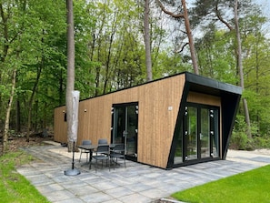 A modern wooden cabin with a glass enclosure, a stone patio, and a table with chairs. - Just Nature Light 4 (Arnhem)