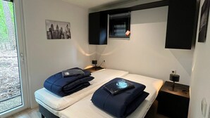 A modern hotel room with a bed, two bedside tables, a window with blinds, a wall-mounted TV, and a cityscape picture. - Just Nature Light 4 (Arnhem)