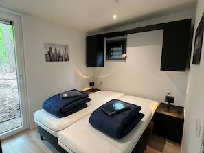 A modern hotel room with a bed, two bedside tables, a window with blinds, a wall-mounted TV, and a cityscape picture. - Just Nature Light 4 (Arnhem)