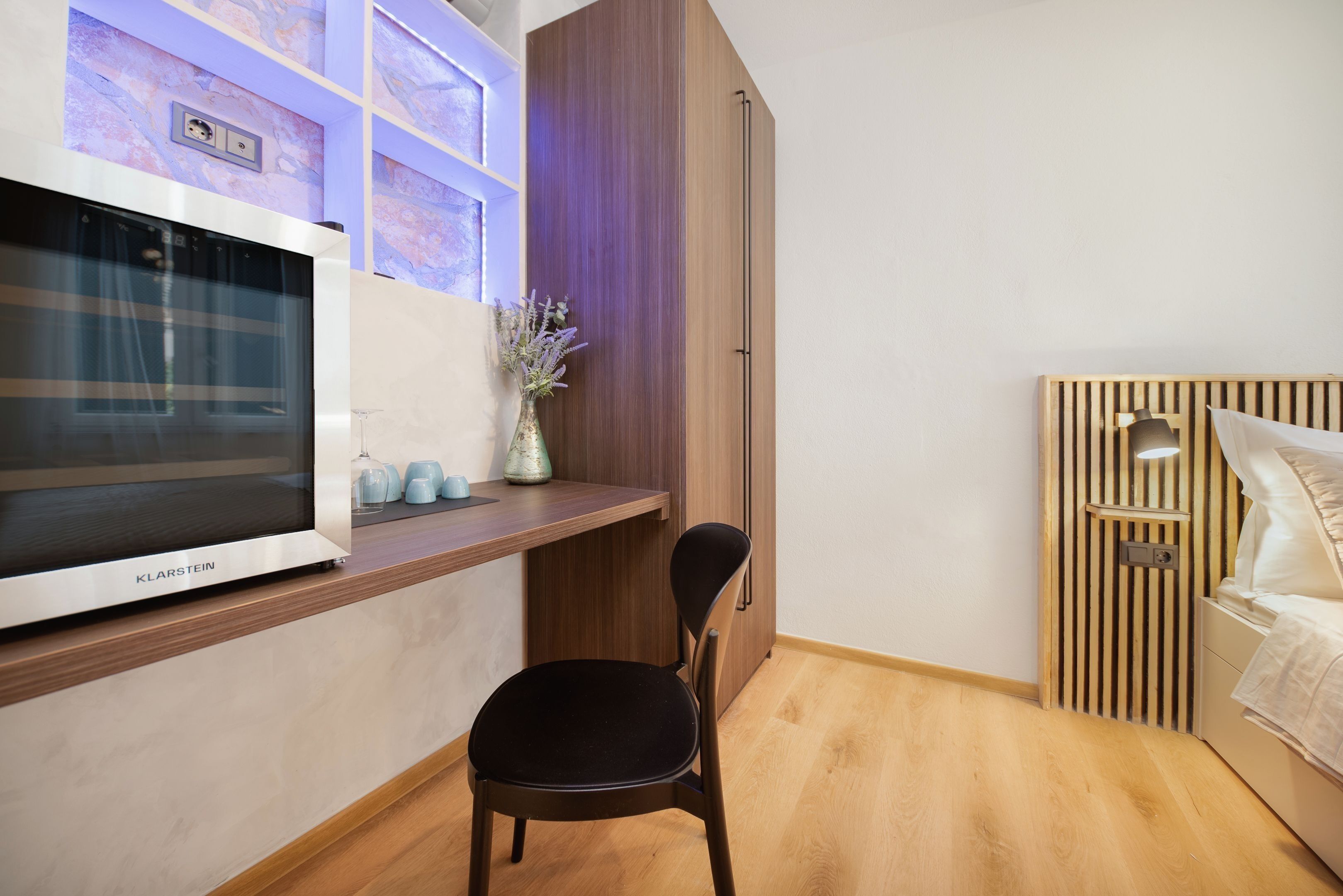 A modern room with a wooden floor, a black chair, a bed with a wooden headboard, a TV on a wooden shelf, and a vase with flowers.