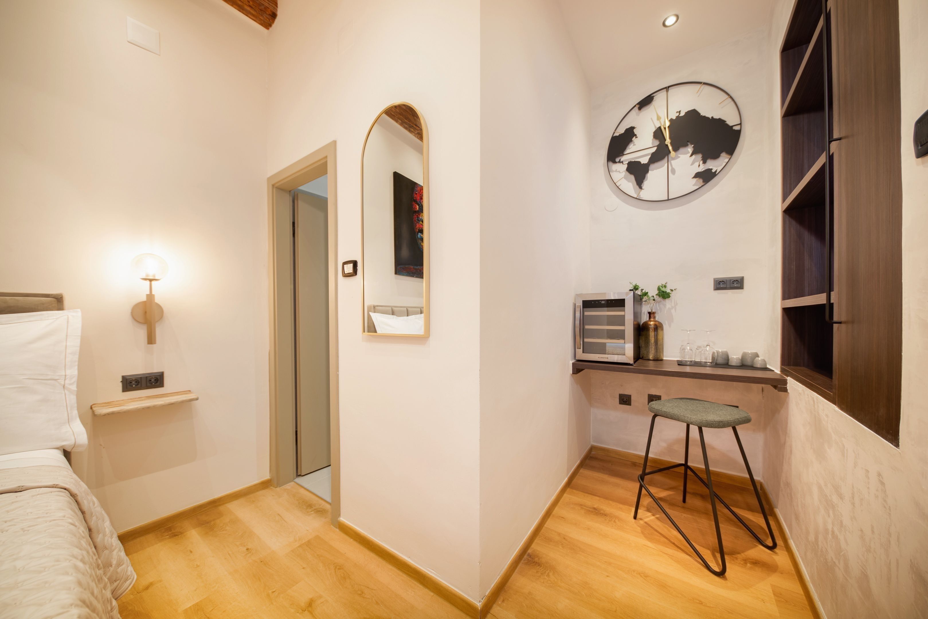 A bedroom with a bed, a bedside table, a mirror, a wall clock, and a small stool.