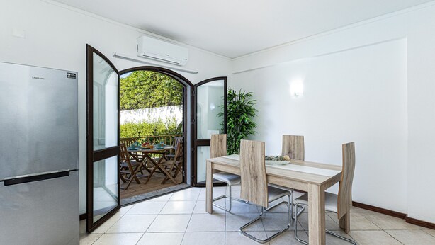 Eine moderne Küche mit einem Edelstahlkühlschrank, einem Holztisch mit Stühlen und einer offenen Tür zu einem Balkon mit Sitzmöbeln.