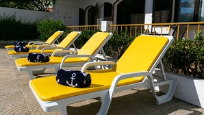 Eine Reihe gelber Liegestühle mit weißen Gestellen und Kissen, einige mit Strandtüchern im Segler-Design, auf einer gepflasterten Terrasse mit einem Gebäude im Hintergrund.