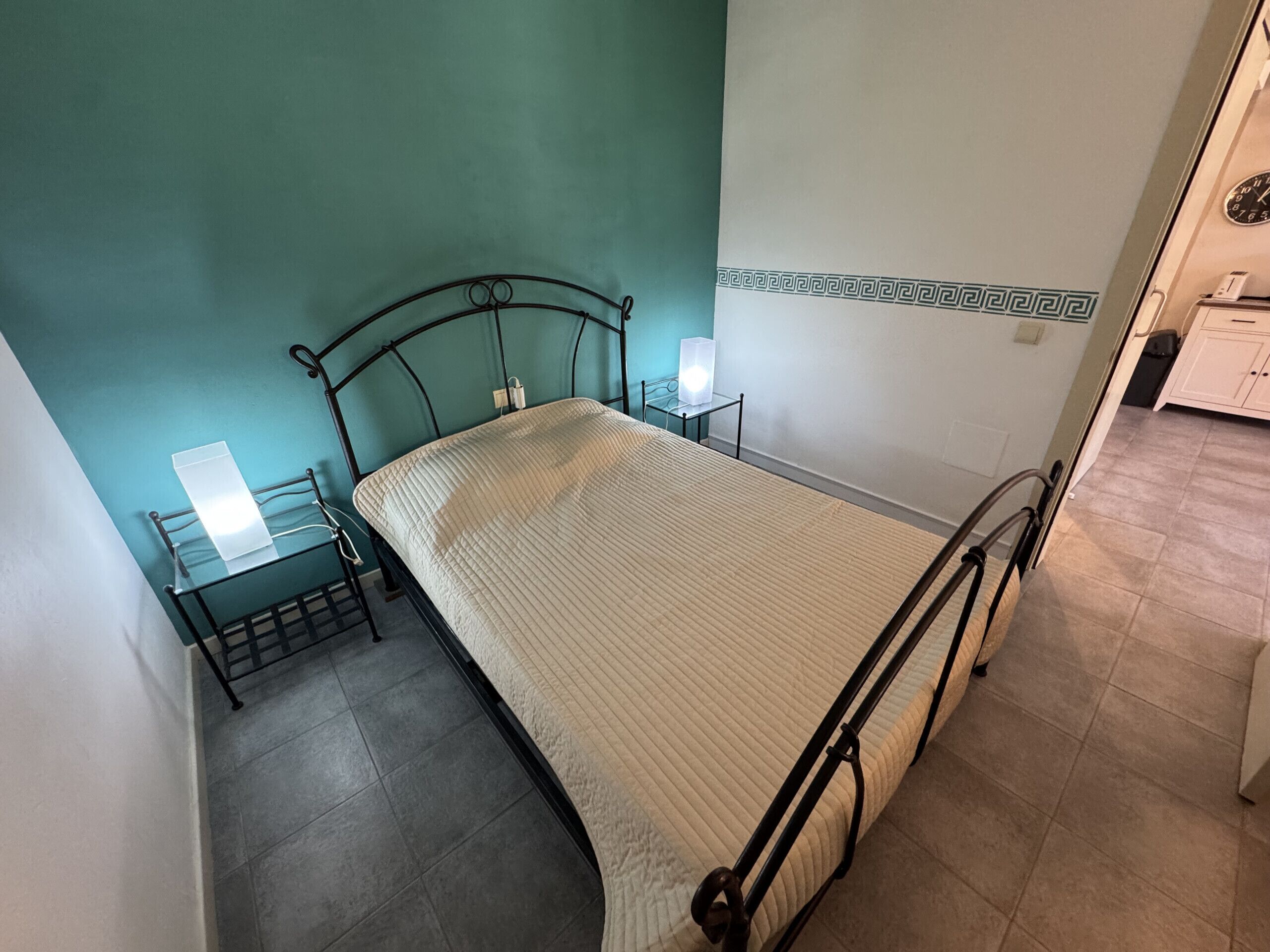 Un lit simple avec un cadre en métal, une table de chevet avec une lampe et une horloge fixée au mur.