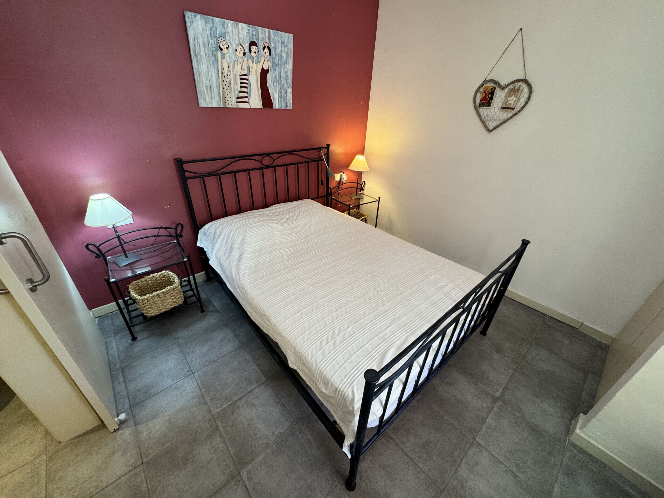 Un lit simple avec un cadre en métal, une table de chevet avec une lampe, une œuvre d’art accrochée au mur et une décoration en forme de cœur.