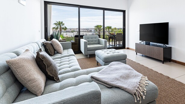 Ein modernes Wohnzimmer mit einem grauen Ecksofa, einem grauen Sessel, einem Fernseher auf einem Holzschrank und einem großen Fenster mit Blick auf die Stadt.