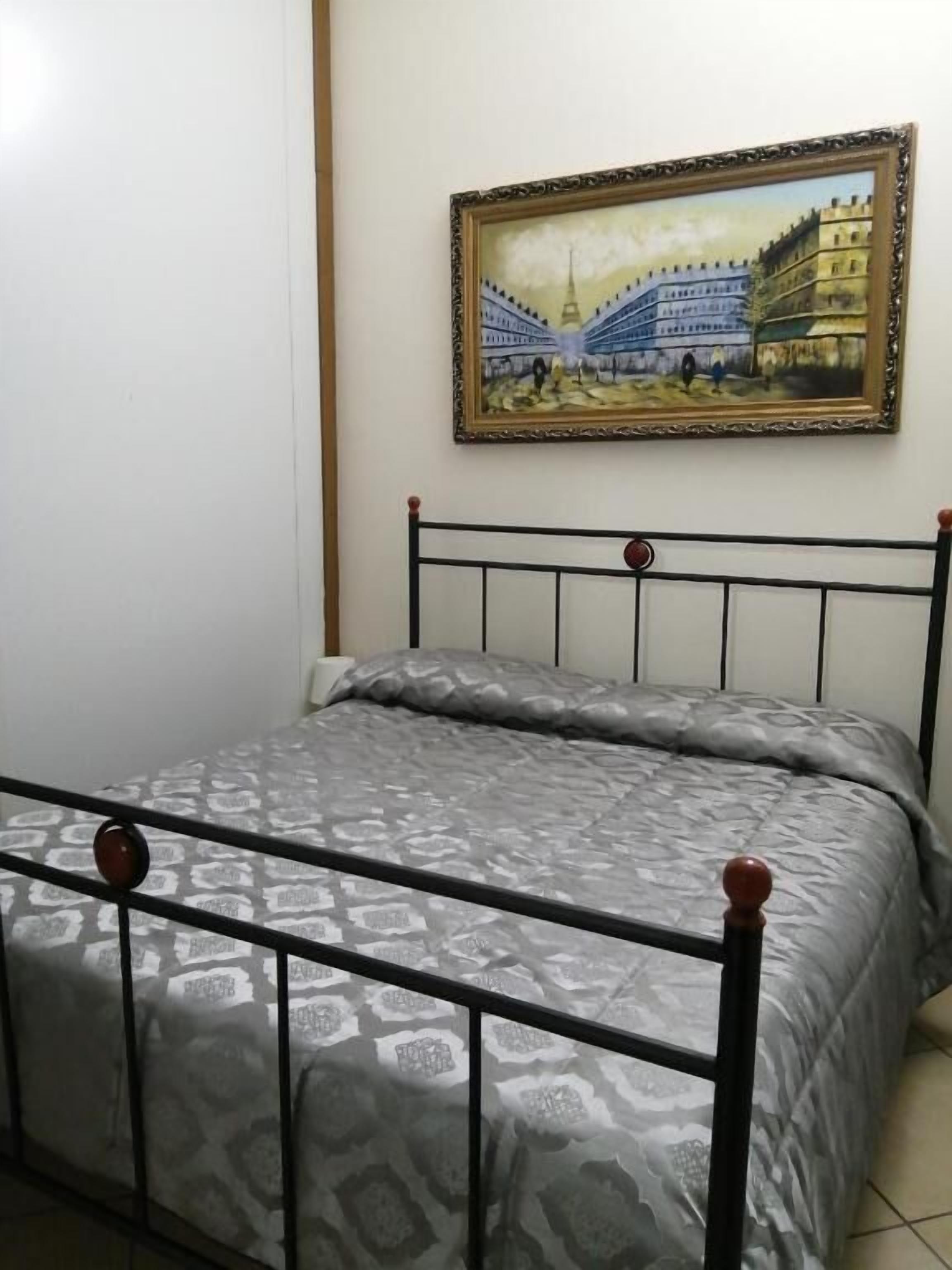 Una cama con estructura metálica y ropa de cama estampada, un cuadro colgado en la pared que muestra un edificio, y una pared lisa.