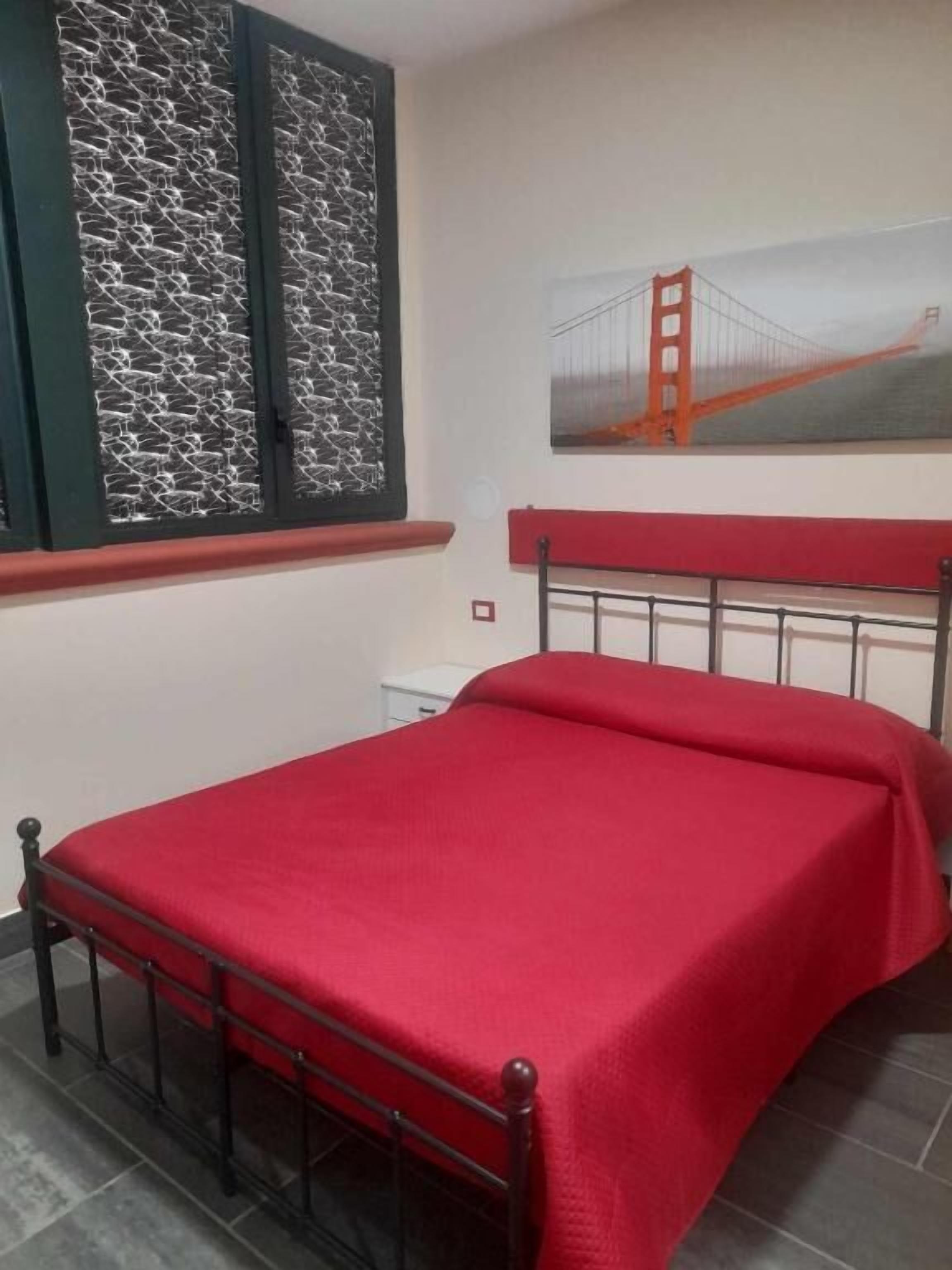 A bedroom with a metal bed frame, a red bedspread, a window with a decorative screen, and a painting of the Golden Gate Bridge on the wall.