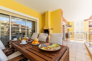Ein Balkon mit Holztisch, Stühlen und einer Frühstücksvorrichtung, darunter Obst und eine Schüssel.