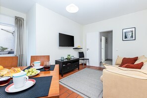 Ein modernes Hotelzimmer mit Flachbildfernseher, einer Essecke mit Frühstückstisch, einer Couch und Blick ins Freie.