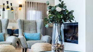 Ett vardagsrum med ett glasbord, en vas med vita blommor och en öppen spis i bakgrunden.