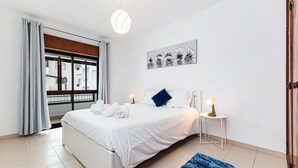 A bedroom with a bed, a nightstand, a lamp, a window with curtains, a painting on the wall, and a blue rug. - Cozy Place Lagos Apartment | Beach | Couples & Nom (Lagos)