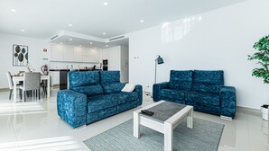 Ein modernes Wohnzimmer mit zwei blauen Sofas, einem Couchtisch, einer Essecke mit Tisch und Stühlen sowie einer Küche im Hintergrund.