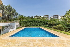 Ein rechteckiger Swimmingpool mit einer gefliesten Umrandung, umgeben von einer angelegten Grünfläche mit einer Hecke und einem Gebäude im Hintergrund.