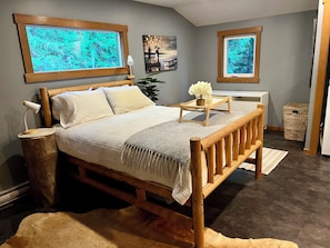 A bedroom with a wooden bed, a nightstand, a lamp, a small table with a candle and flowers, a picture on the wall, and a window with a view of trees.