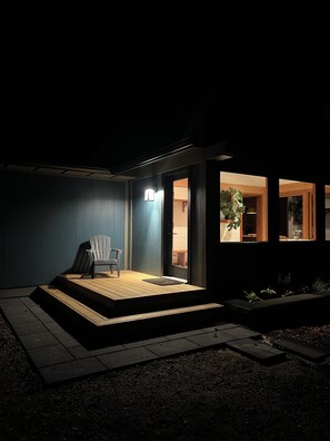 Un espacio exterior oscuro con una terraza de madera, una sola silla y una puerta que da a un espacio interior.