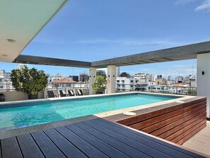 Ein Pool auf dem Dach mit Blick auf die Stadtlandschaft, eine Holzterrasse und Liegestühle.