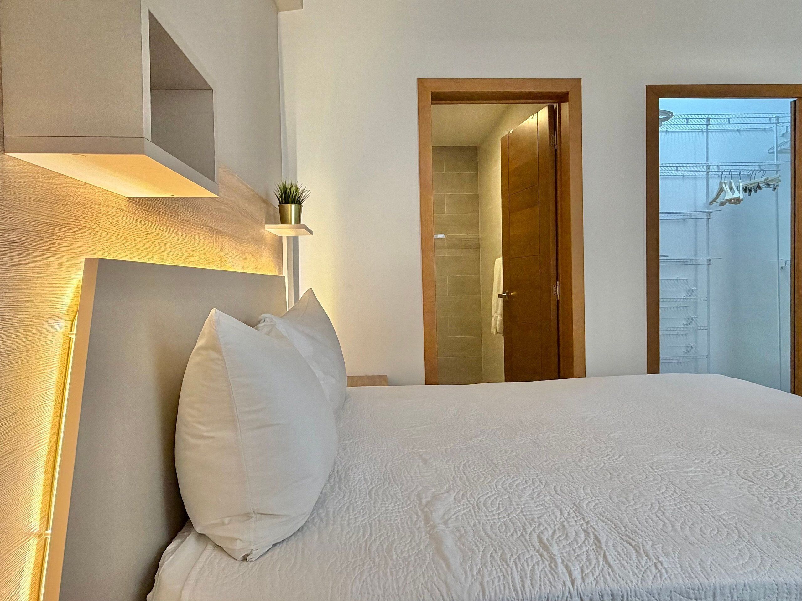 A bedroom with a bed, a bedside table, a wall-mounted shelf with a plant, and a door leading to a balcony.