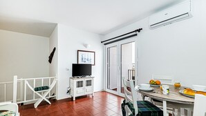 Una habitación con una cama blanca, un televisor sobre un mueble blanco, una mesa de comedor con sillas y un ventanal corredizo que da al exterior.