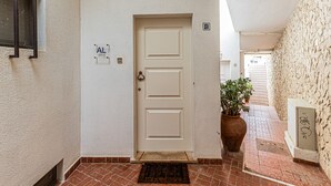 Un pasillo con una puerta blanca marcada con las señas 'B' y 'AL', una planta en maceta y un piso de baldosas.