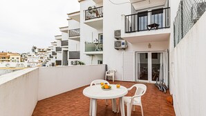 Un balcón con una mesa y sillas blancas, una pequeña mesa con fruta y una bebida, y vista del paisaje urbano.
