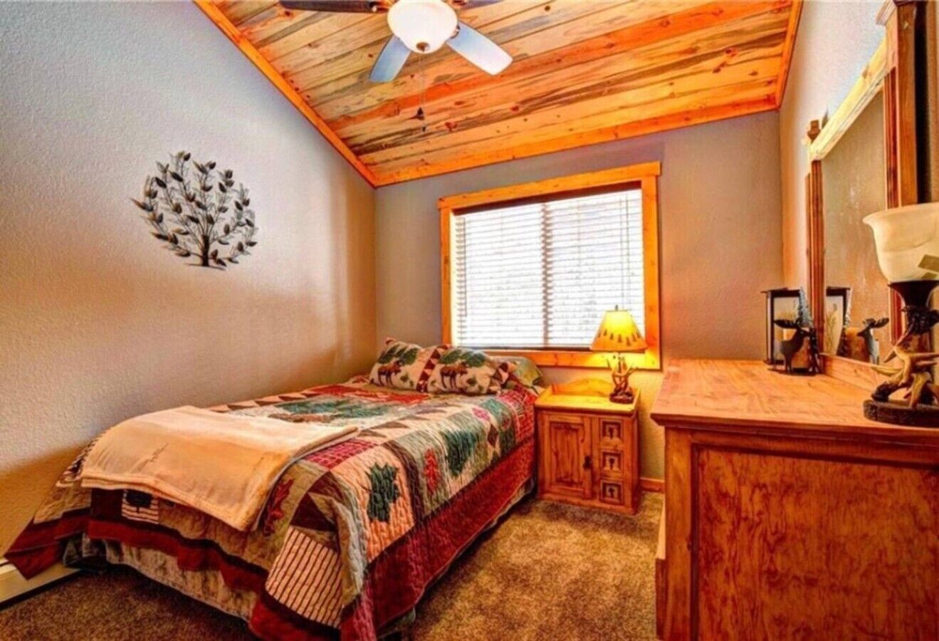 Een slaapkamer met een bed, nachtkastje, lamp, dressoir, een raam met jaloezieën en een plafondventilator.