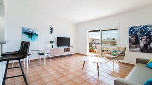 Ein modernes Wohnzimmer mit Fernseher, weißen Möbeln, einem Balkon mit Aussicht und Kunstwerken an den Wänden.