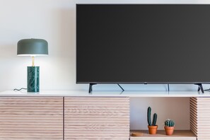A flat-screen TV mounted on a white wall, a marble lamp on a wooden cabinet, and two potted cacti on a shelf. - Casa da Galé: Near Meia Praia | Perfect place w/ S (Lagos)
