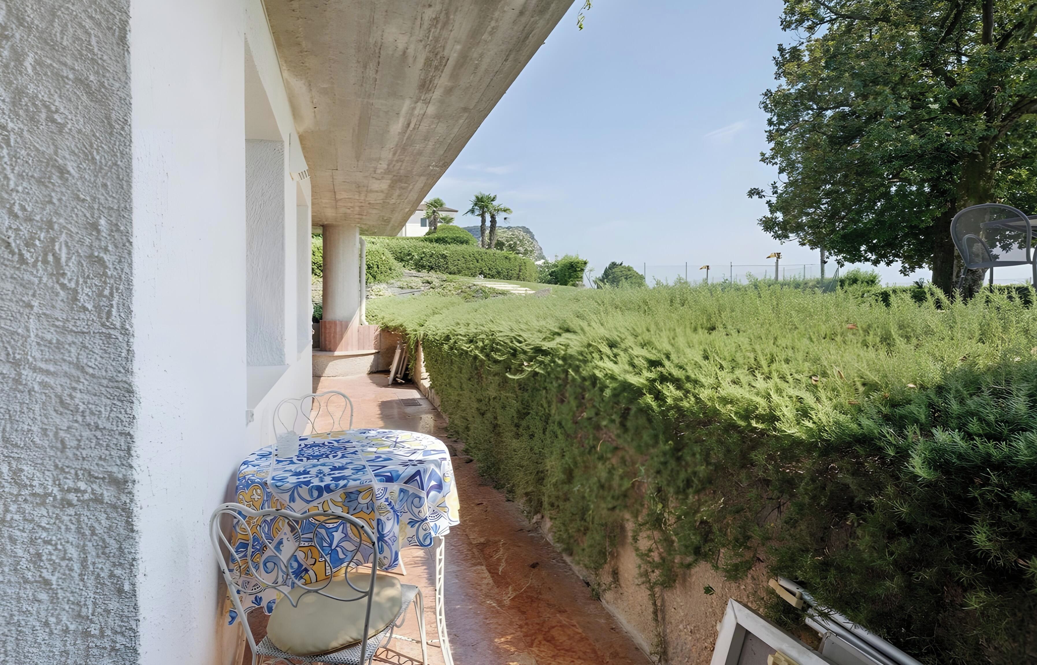 A covered outdoor seating area with a table and chairs, a concrete structure overhead, and a view of a landscaped garden with a hedge and trees.