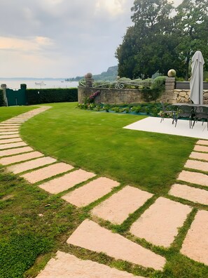Ein angelegter Garten mit einem Steinweg, einer Sitzgelegenheit mit Stühlen und einem Tisch sowie Blick auf ein Gewässer.